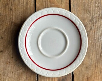 May include: A white ceramic saucer featuring a central circular indentation for a cup. The rim is decorated with a raised scalloped pattern and a single thin red circular line that encircles the center, creating a clean and simple design.