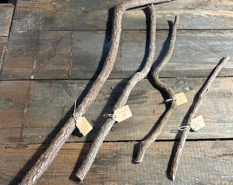 May include: Four rustic, natural tree branches of varying lengths and shapes, arranged on a weathered wooden surface. Each branch has a textured, brown bark and is tagged with a small, blank price tag. The branches are ideal for crafting or decorative purposes.
