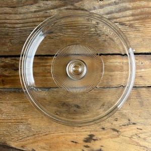 May include: A clear glass lid with a circular design. The lid features a raised, ridged center and a rounded edge. The glass is transparent, allowing a view of the wooden surface beneath. A kitchenware item.