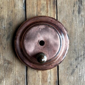 May include: A round copper-colored metal object with a small knob on the bottom and a hole in the center. The object is on a wooden surface.