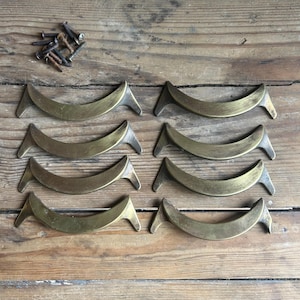 May include: Eight antique brass drawer pulls with a curved design and pointed ends are arranged on a wooden surface. A small pile of screws is visible in the upper left corner. The pulls have a vintage aesthetic.