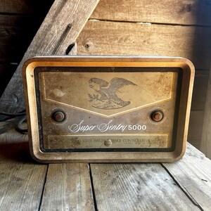 May include: Vintage Super Sentry 5000 electric fence controller. The rectangular device has a gold-colored face with an eagle emblem and the words "Super Sentry 5000". Two round knobs are visible. The frame is a dark brown color.