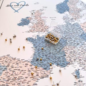 Peut inclure: Une carte détaillée de l'Europe avec des punaises indiquant des lieux. La carte est de couleur bleue et beige, avec le texte "Steve and Carols ADVENTURES" en haut. Un récipient en plastique transparent contient des punaises supplémentaires.