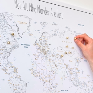 Peut inclure: Une carte du monde murale blanche avec le texte Not All Who Wander Are Lost en haut. La carte affiche les continents et les pays en contours noirs avec des accents dorés. Plusieurs punaises dorées sont insérées à divers endroits.