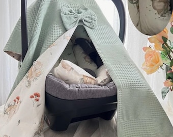 May include: A baby car seat cover in a soft green waffle weave fabric, adorned with a matching bow. The cover features a white and light green patterned fabric with koala bear illustrations and is trimmed with white pom-poms. The car seat is gray.
