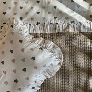 Puede incluir: Juego de cama blanco con estampado de corazones grises y beige. El juego incluye una almohada con borde con volantes y una sábana a juego con ribete con volantes. El fondo es una tela beige texturizada.