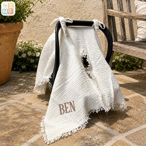 May include: A cream-colored baby car seat cover with the name "BEN" embroidered in brown. The cover has a fringed edge and is draped over a black car seat handle. The background includes a terracotta pot with a plant and a wooden bench.