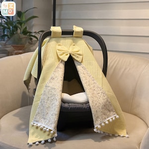Könnte beinhalten: Eine gelbe Baby-Autositzabdeckung mit Schleife und floralem Besatz. Die Abdeckung besteht aus einem strukturierten Stoff und hat weiße Pompons an den Rändern. Die Abdeckung ist für einen Autositz konzipiert.