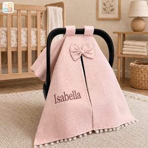 May include: A light pink waffle-textured car seat cover featuring a central bow at the top and the name Isabella embroidered in a darker pink thread. The cover has a split front design and is finished with a row of small white pom-pom trim.