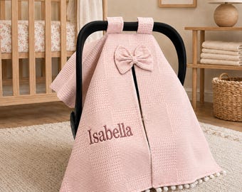 May include: A light pink waffle-textured car seat cover featuring a central bow at the top and the name Isabella embroidered in a darker pink thread. The cover has a split front design and is finished with a row of small white pom-pom trim.