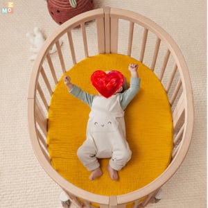 May include: An oval wooden cot with vertical slats and a mustard yellow fitted sheet. Inside, a person wears a light green long-sleeved shirt under a beige romper featuring a simple embroidered animal face on the front chest area.