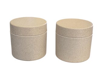 May include: Two beige, cylindrical containers with matching lids. The containers have a speckled texture and a simple, modern design. The lids fit snugly on top, creating a clean, minimalist look.