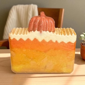 May include: A rectangular bar of soap with layers of yellow, orange, and white, topped with a small orange pumpkin. The soap is sitting on a wooden surface. The soap is likely handmade and suitable for fall or autumn decor.