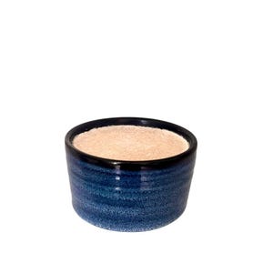 May include: A small, round, blue ceramic bowl with a black rim. The bowl has a textured, painted finish and contains a light brown substance. The bowl is set against a white background.