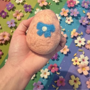 May include: A peach-colored, egg-shaped felt ornament with a blue bow and white center. The ornament is held in a hand, surrounded by colorful felt flowers. The felt egg is a decorative item.