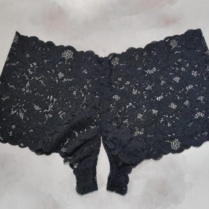 May include: Black lace underwear with a floral pattern. The garment has a boy short style with a scalloped edge along the waist and leg openings. The fabric is semi-sheer, revealing the background.