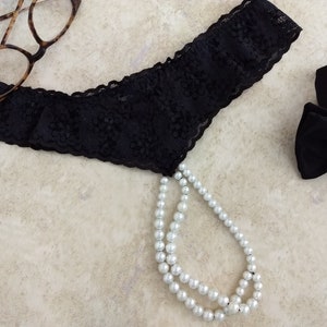 May include: Black lace lingerie with a pearl necklace attached. The lingerie is on a light beige surface.