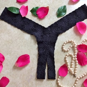 May include: Black lace underwear with a cutout detail. The underwear is lying on a light brown surface with pink rose petals and a pearl necklace.