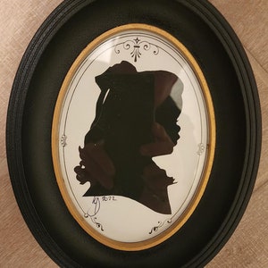 May include: A black silhouette of a person in a hat, framed in a black oval frame with a gold inner border. The silhouette is on a white background with a handwritten signature and the date "2012".