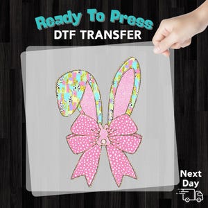 May include: A colorful DTF transfer featuring bunny ears and a bow. The ears have a pink center and are outlined in gold, with a patterned design. The bow is pink with white polka dots. The text "Ready To Press" and "DTF TRANSFER" is at the top.