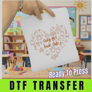 May include: A white DTF transfer sheet featuring a brown line art design in the shape of a heart. The design includes illustrations of books, pumpkins, mugs, mittens, and hats, with the text "cozy girl book club" centered inside the heart.