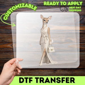 Może przedstawiać: Arkusz transferowy DTF z ilustracją stojącego jelenia, ubranego w bezrękawową, kremową sukienkę kopertową z wiązaniem w talii, trzymającego małą, pasującą kremową torebkę. Jeleń ma jasnobrązowe futro i duże uszy.