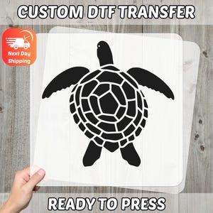 May include: Black sea turtle graphic on a clear transfer sheet. The text "CUSTOM DTF TRANSFER" and "READY TO PRESS" are displayed. The image is held over a wooden surface.