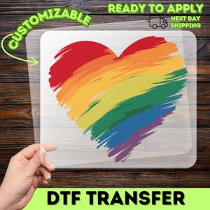 May include: A clear DTF transfer sheet featuring a heart graphic composed of horizontal brushstrokes in rainbow colors including red, orange, yellow, green, blue, and purple. The design is printed on a transparent film with a smooth finish.