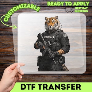 May include: A DTF transfer sheet featuring a tiger with a human-like physique dressed in black tactical gear. The figure wears a vest labeled SECURITY, carries a rifle, and has a tail visible. The design includes a US flag patch on the shoulder.