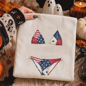 May include: A white sweatshirt featuring a graphic print of a two-piece bikini. The bikini top and bottom are decorated with a patchwork pattern of red and white stripes and blue fabric with white stars, mimicking the American flag design.
