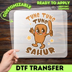 May include: A DTF transfer featuring a cartoon character with the text "Tung Tung Sahur". The character is brown with a smiling face, holding a wooden bat, and surrounded by stars. The words "Customizable" and "Ready to Apply" are also present.