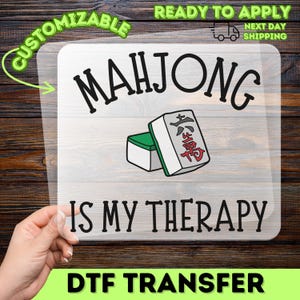 May include: A clear DTF transfer with the words "MAHJONG IS MY THERAPY" in black. The image includes a graphic of two Mahjong tiles. The transfer is labeled "CUSTOMIZABLE" and "READY TO APPLY".