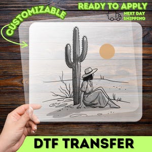 May include: A clear DTF transfer sheet featuring a black and white line art illustration of a person wearing a hat and dress sitting in the desert next to a tall saguaro cactus under a solid tan circle representing the sun in the sky.