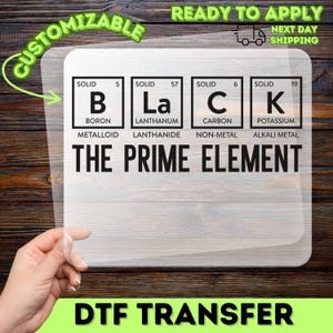 May include: A clear DTF transfer with the word "BLACK" spelled out using periodic table elements. The text "THE PRIME ELEMENT" is below the word "BLACK". The transfer is on a wooden background. The words "CUSTOMIZABLE" and "READY TO APPLY" are also visible.