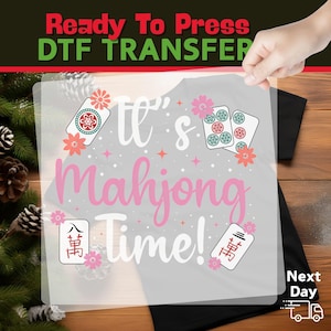 May include: A clear DTF transfer sheet with the text "It's Mahjong Time!" in pink and white, along with decorative mahjong tiles and flowers. The words "Ready To Press" are at the top in red and green.