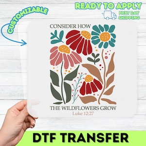 May include: A white DTF transfer with a floral design in red, pink, and teal. The text reads "CONSIDER HOW THE WILDFLOWERS GROW Luke 12:27". The transfer is labelled "DTF TRANSFER" and "CUSTOMIZABLE".