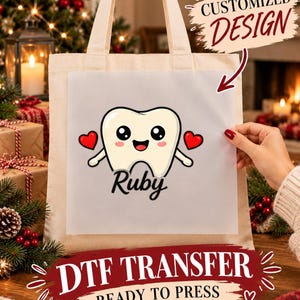 Puede incluir: Una bolsa de tela beige con una transferencia DTF que muestra un diente de dibujos animados, dos corazones rojos y el nombre Ruby en letra cursiva negra. La transferencia incluye el texto CUSTOMIZED DESIGN y DTF TRANSFER READY TO PRESS.