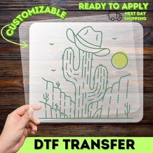 May include: A clear DTF transfer sheet featuring a green line art illustration of a saguaro cactus wearing a cowboy hat in a desert landscape with a sun and birds. The design includes the text CUSTOMIZABLE, READY TO APPLY, and NEXT DAY SHIPPING.