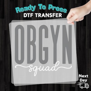 May include: A gray DTF transfer with the text "Ready To Press" and "OBGYN squad" in a script font. The transfer is held up by a hand against a dark wood grain background.