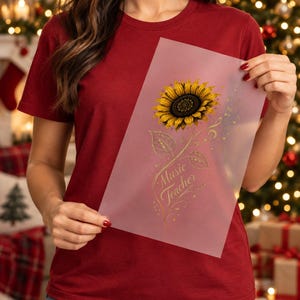 May include: A maroon short-sleeved t-shirt and a translucent sheet featuring a yellow sunflower illustration with gold musical notes and the text Music Teacher in a cursive font, with a dark center and detailed green leaves on the stem.