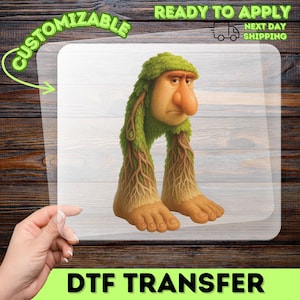 May include: A DTF transfer featuring a whimsical cartoon character with a large nose, green leafy hair, and legs resembling tree roots. The text "CUSTOMIZABLE" and "READY TO APPLY" are visible. The image is on a clear background.