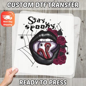 May include: A DTF transfer sheet with a design of a black-lipped mouth, a red snake, and the words "Stay Spooky." The design includes spiders, a spiderweb, and red roses. The text "READY TO PRESS" is also visible.