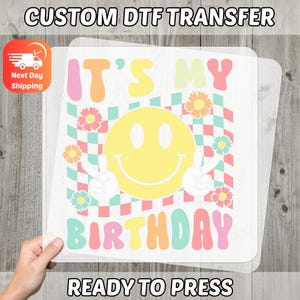 May include: A ready-to-press DTF transfer featuring a large yellow smiley face with peace signs, surrounded by flowers and a checkered background. The text reads "IT'S MY BIRTHDAY" in colorful letters. The image also includes the text "CUSTOM DTF TRANSFER" and "READY TO PRESS".