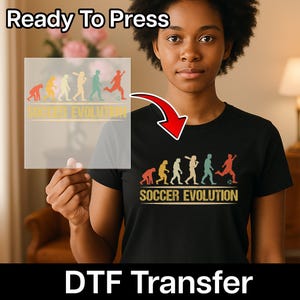 May include: Black t-shirt featuring a "Soccer Evolution" design in retro colours. The graphic depicts a sequence of figures playing football. The words "Ready To Press" and "DTF Transfer" are also visible.