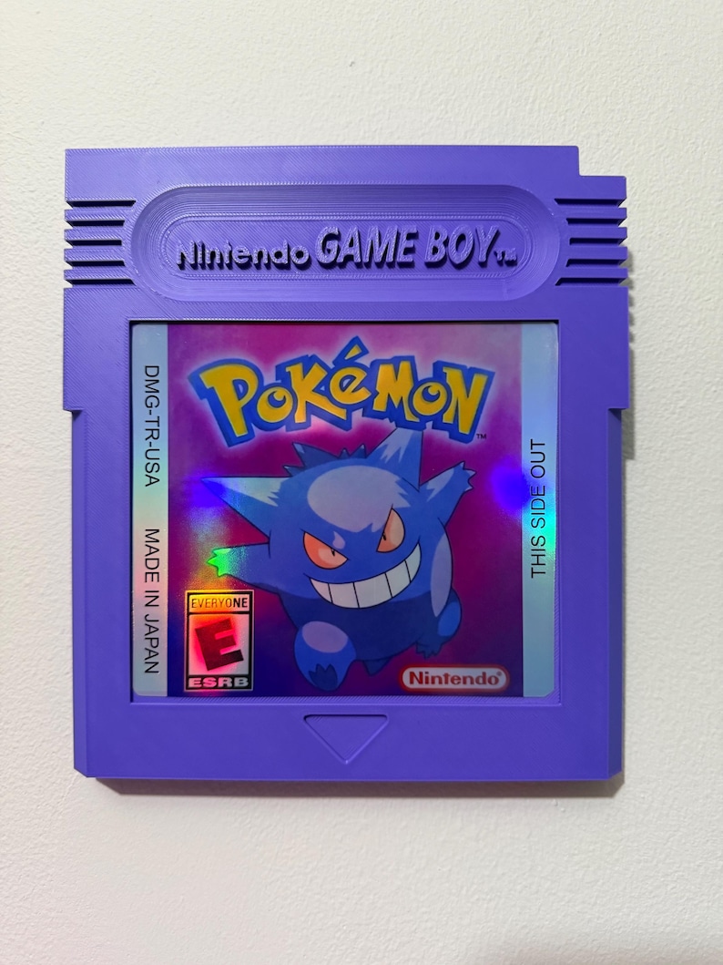 May include: A purple Nintendo Game Boy cartridge wall display featuring a holographic Pok&eacute;mon label with Gengar, the text Pok&eacute;mon, Nintendo GAME BOY, DMG-TR-USA, MADE IN JAPAN, THIS SIDE OUT, and an ESRB Everyone E rating symbol.