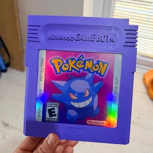 May include: A purple Nintendo Game Boy video game cartridge featuring a holographic label with the Pok&eacute;mon logo and an illustration of the character Gengar. The label includes the text DMG-TR-USA, THIS SIDE OUT, MADE IN JAPAN, and an ESRB E rating.