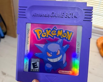 Puede incluir: Un cartucho de videojuego Nintendo Game Boy de color morado con una etiqueta holográfica que muestra el logotipo de Pokémon y una ilustración del personaje Gengar. La etiqueta incluye el texto DMG-TR-USA, THIS SIDE OUT, MADE IN JAPAN y ESRB E.