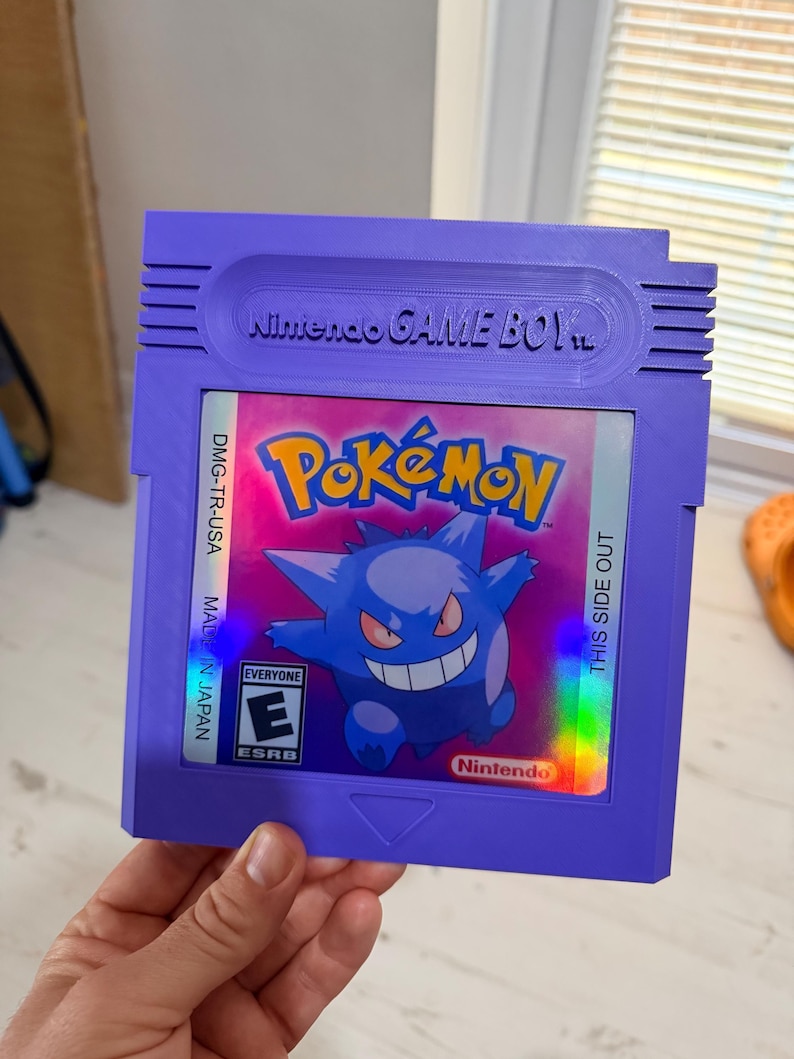 May include: A purple Nintendo Game Boy video game cartridge featuring a holographic label with the Pok&eacute;mon logo and an illustration of the character Gengar. The label includes the text DMG-TR-USA, THIS SIDE OUT, MADE IN JAPAN, and an ESRB E rating.