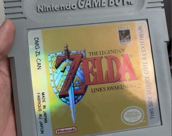 May include: A gray Nintendo Game Boy video game cartridge for The Legend of Zelda: Link's Awakening. The label features a gold background, a blue shield with a sword, and the game title in red text. It includes the Nintendo logo and DMG-ZL-CAN.