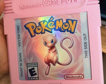 Puede incluir: Un cartucho de videojuego Nintendo Game Boy de color rosa claro con una etiqueta holográfica que muestra el logotipo de Pokémon, una ilustración del personaje Mew, el símbolo de clasificación ESRB Everyone y el texto DMG-WS-USA, MADE IN JAPAN, THIS SIDE OUT.